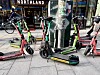 Flere utleide elsparkesykler står tett på et fortau i en bygate.