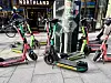Flere utleide elsparkesykler står tett på et fortau i en bygate.