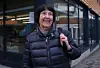 Eldre kvinne med boblejakke smiler utendørs foran butikkfasade i bygate.
