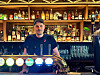 En bartender står bak en opplyst bardisk foran hyller med flasker og glass i en pub.