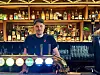En bartender står bak en opplyst bardisk foran hyller med flasker og glass i en pub.