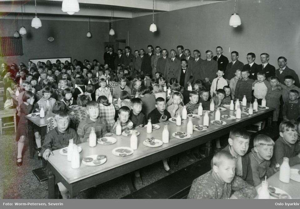 Sagene skole 13. mai 1930, under skolefrokost i matsalen. Elever ved langbord med mat og melkeflasker. Lærere oppstilt langs veggen i bakgrunnen. Forløper til Oslofrokosten. Foto: Severin Worm-Petersen / Oslo byarkiv