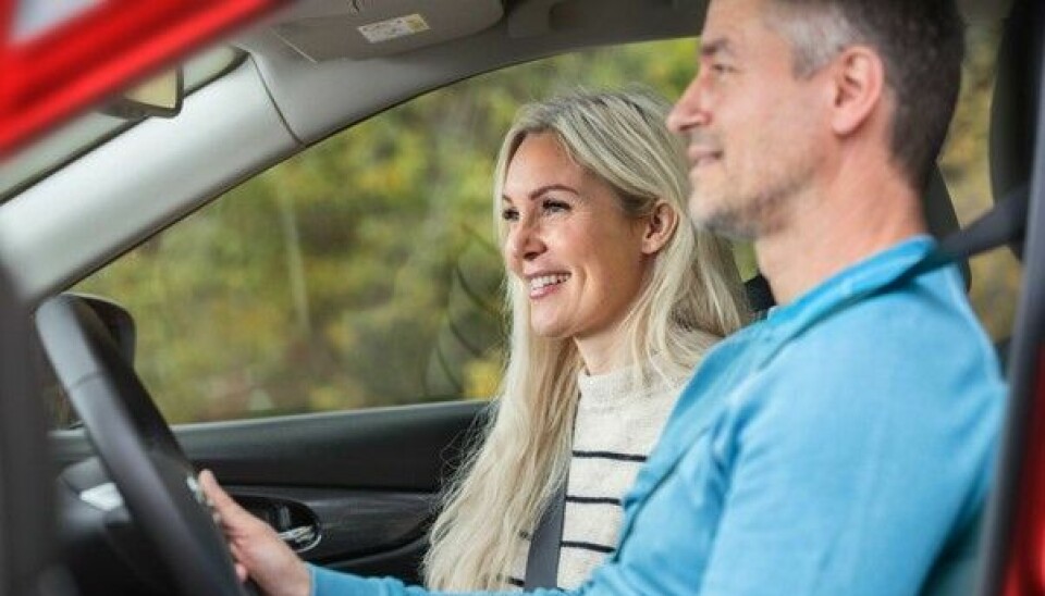 Smilende mannlig sjåfør kjører en bil mens en smilende kvinne sitter i førersetet.