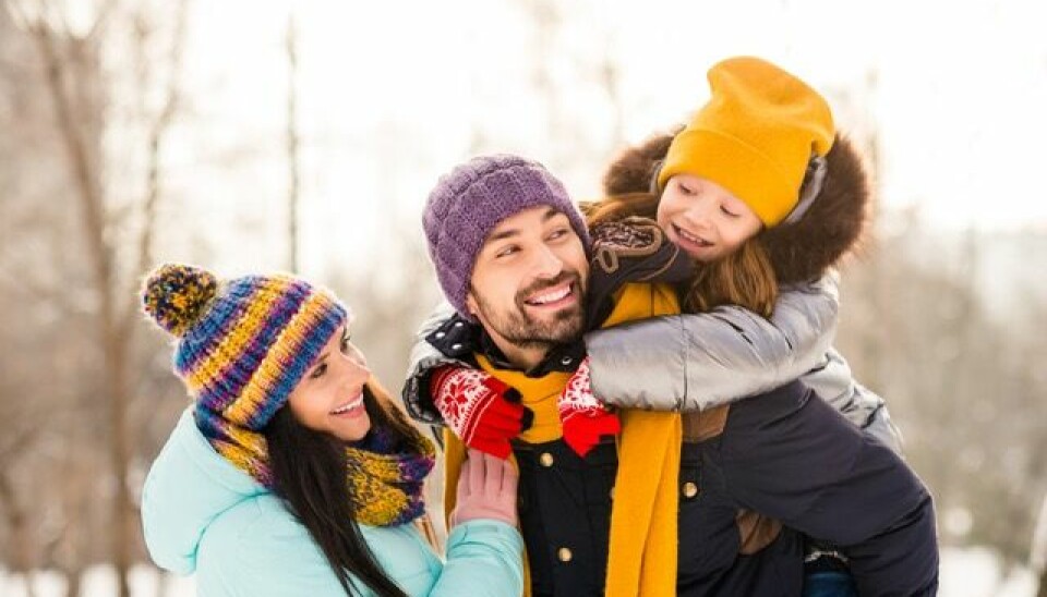 Familie (man, kvinne, ett barn) smiler sammen utendørs en snøfylt vinterdag.