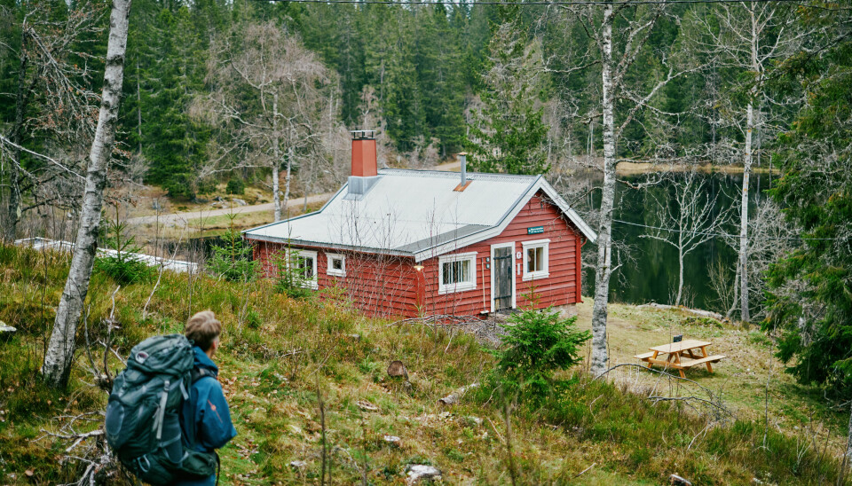 Mann med tursekk ser ned mot en rød hytte med et vann i bakgrunnen.
