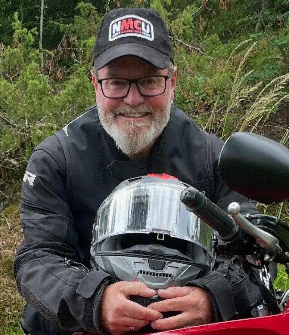 Arild Lind, daglig leder i NMCU, avbildet i motorsykkelklær mens han holder en hjelm. Han smiler. På hodet har han en NMCU-caps.